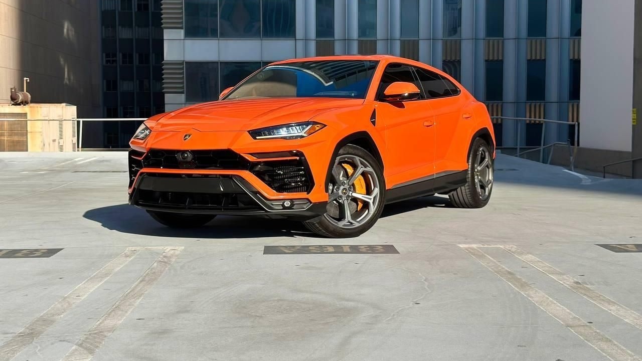 Orange Lamborghini Urus on a rooftop parking deck in front of office towers